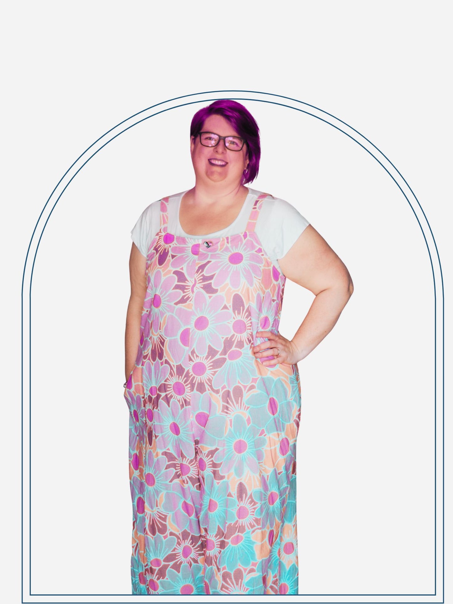 A woman standing in a doorway wearing a square neck jumpsuit with a colorful floral pattern on a purple background, paired with white sandals.