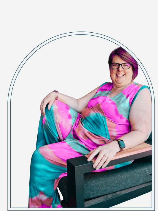 A woman sitting on a chair wearing a colorful tie-dye jumpsuit with a crossover top and elasticated waist, smiling and posing for the camera.