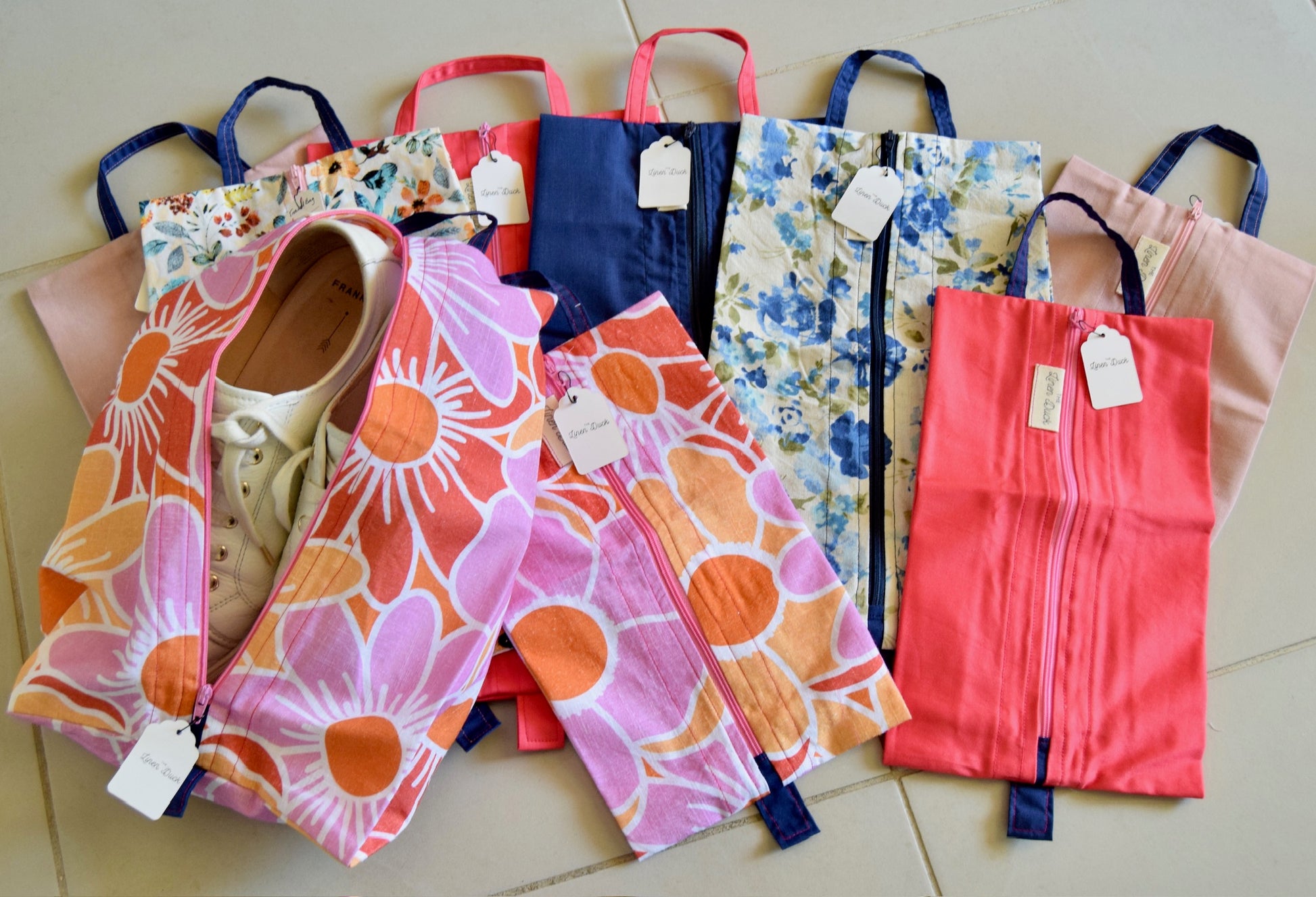 A collection of expandable travel bags in various sizes and colors, featuring a floral pattern on a beige bag, a paisley pattern on an orange bag, and other designs. They are displayed with easy grab handles and tags visible.